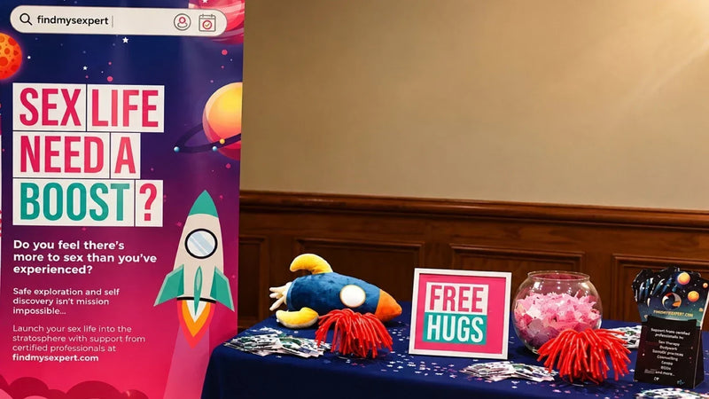 FindMySexpert stand at a London dating event with a colourful “Sex Life Need a Boost?” banner and a Free Hugs sign on the table.