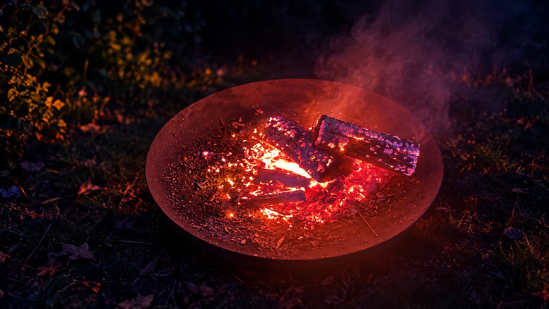 Glowing embers and charred logs in a round fire pit at night, with smoke drifting into the dark garden.