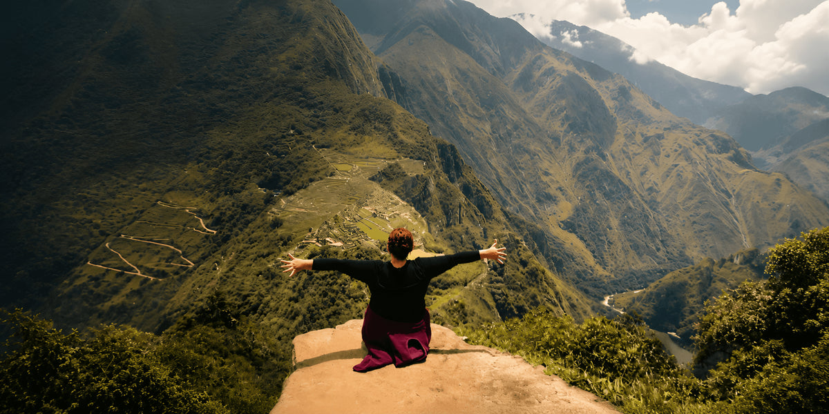 A person sits on a rock in a vast mountain valley with arms outstretched, facing green rugged peaks and clouds. The person is wearing a black top and magenta pants.