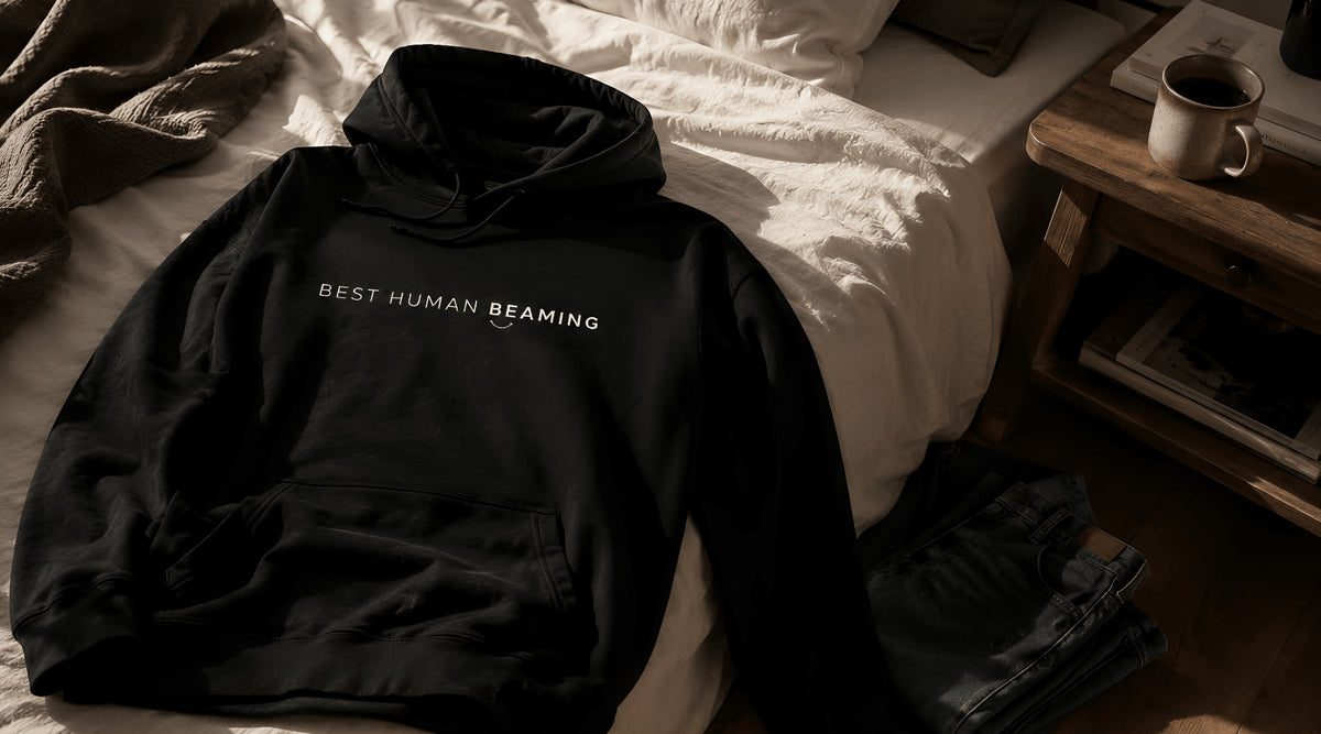 Black hoodie with 'BEST HUMAN BEAMING' text on a bed in a bedroom setting