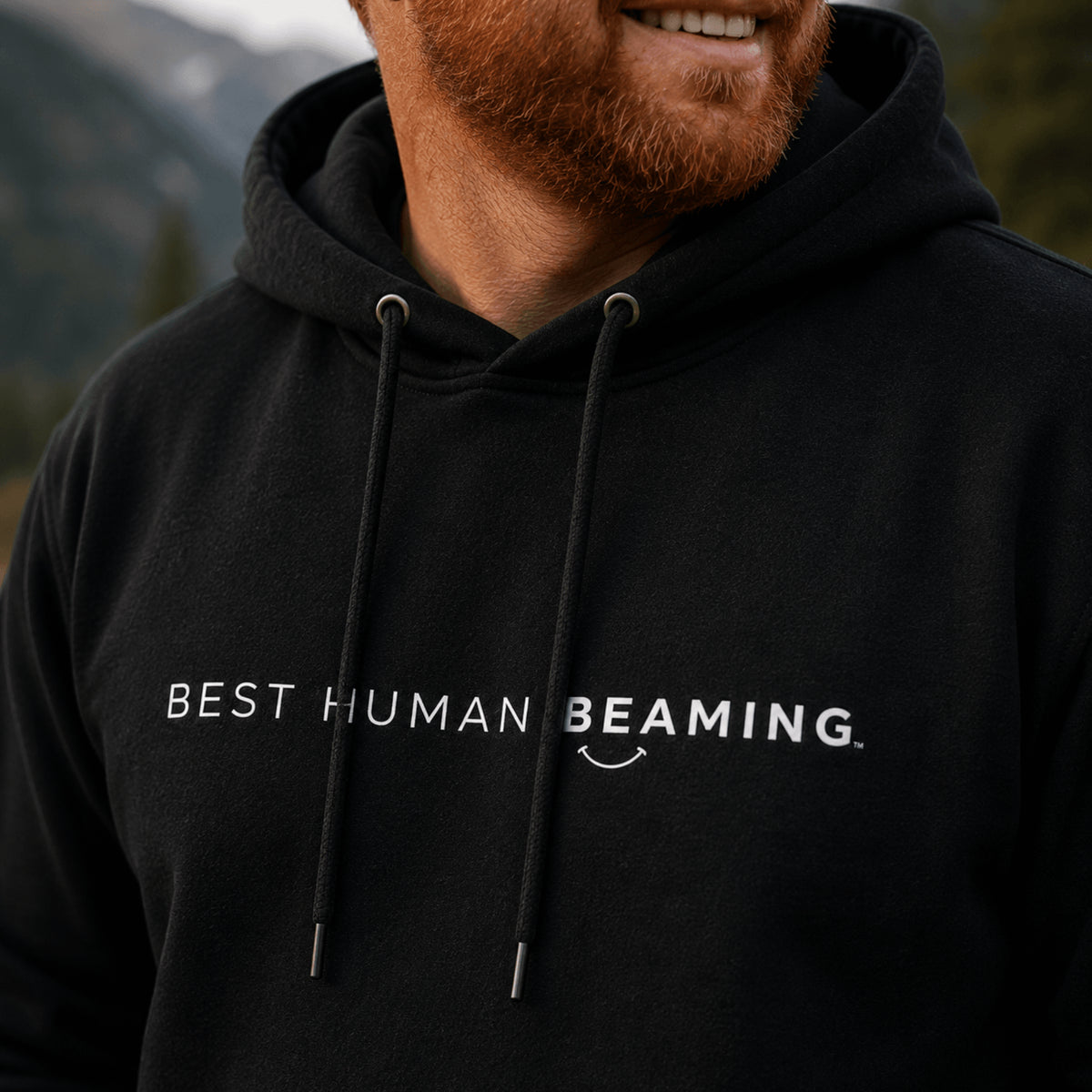 Close-up of the black Best Human Beaming heavyweight hoodie worn by a man, showing the white chest logo, hood and drawcords.