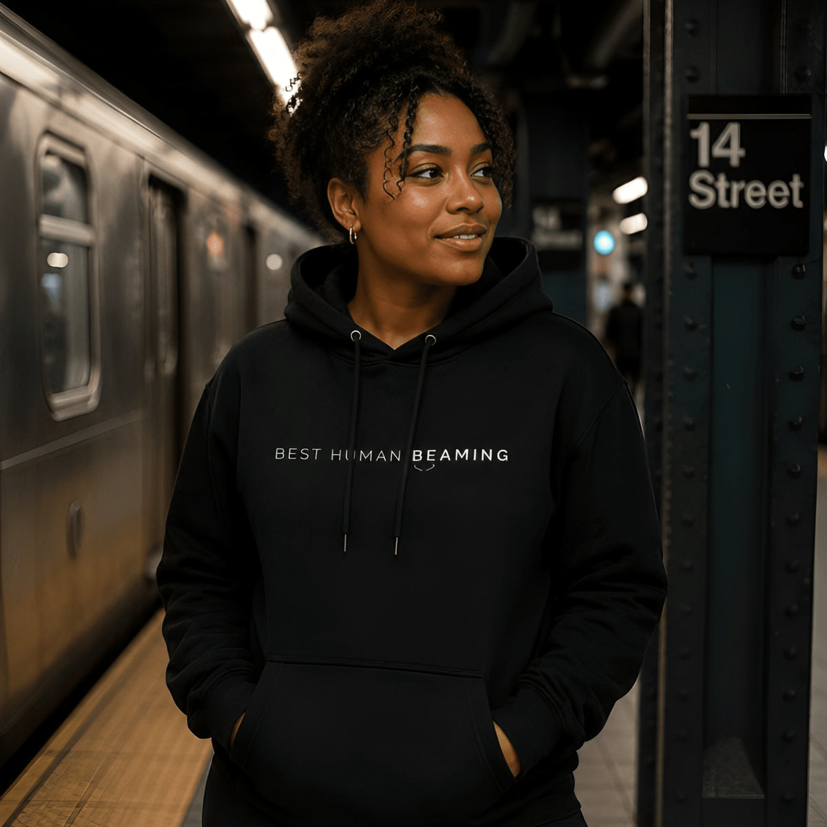 Woman wearing the black Best Human Beaming unisex heavyweight hoodie while standing on a train platform.