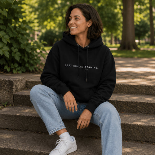 Woman wearing the black Best Human Beaming unisex heavyweight hoodie with jeans while sitting on steps in a park.