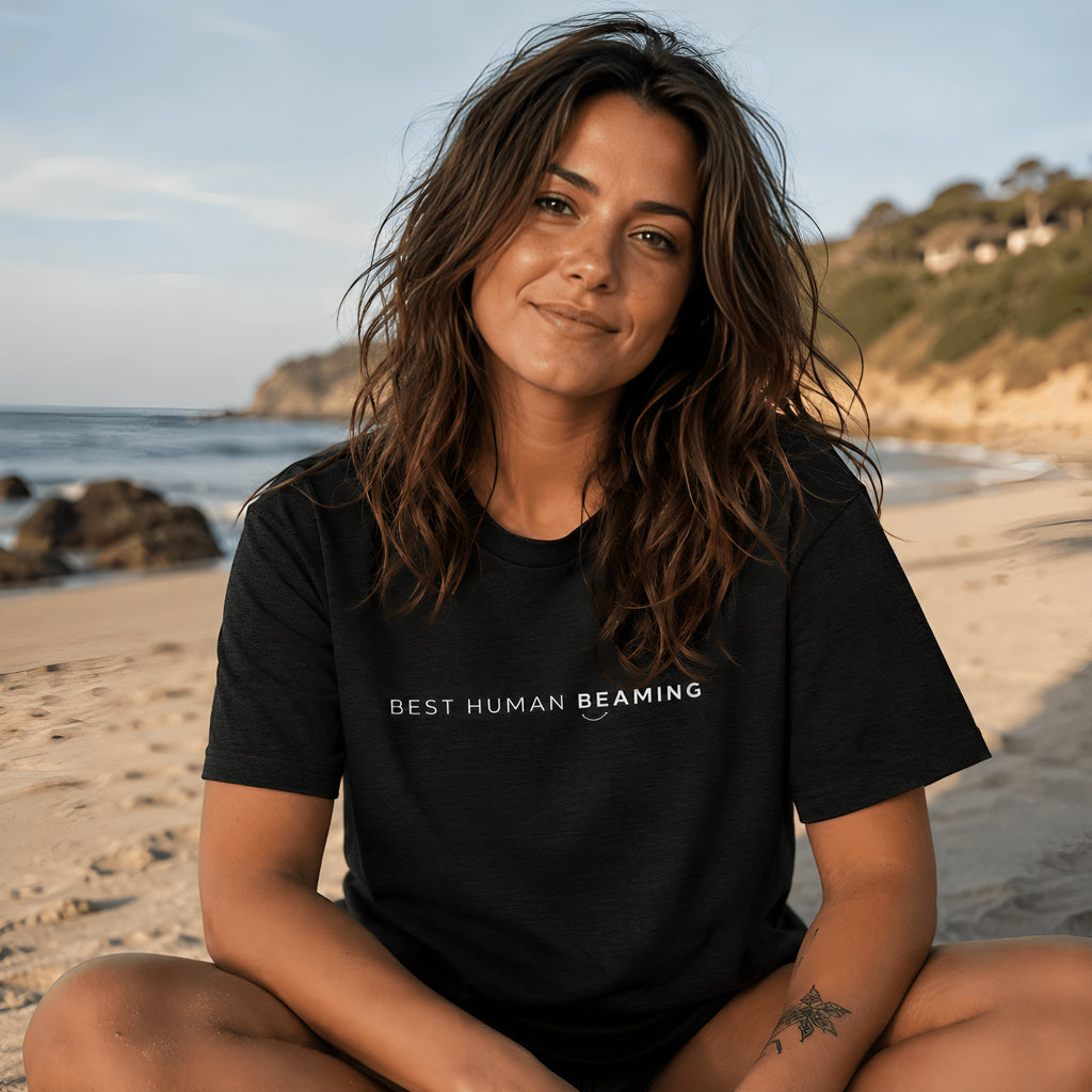 Woman wearing the black Best Human Beaming unisex organic cotton tee while sitting on a beach.