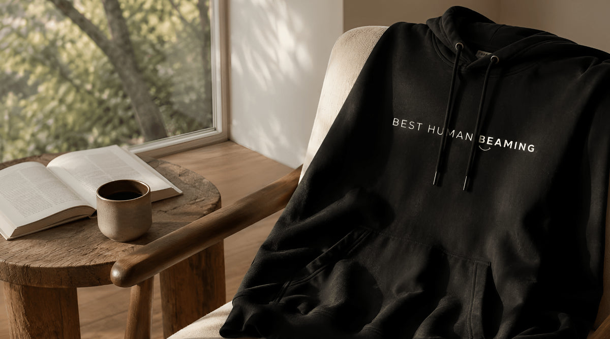 Black hoodie with 'BEST HUMAN BEAMING' text on a chair next to an open book and cup of coffee on a wooden table.