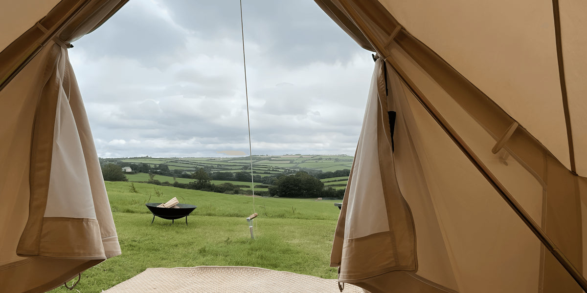 A beige bell tent viewed from inside, with its front flap open to reveal a lush green countryside landscape under a cloudy sky. A small outdoor fire pit and a person sitting on a blanket are visible in the field.