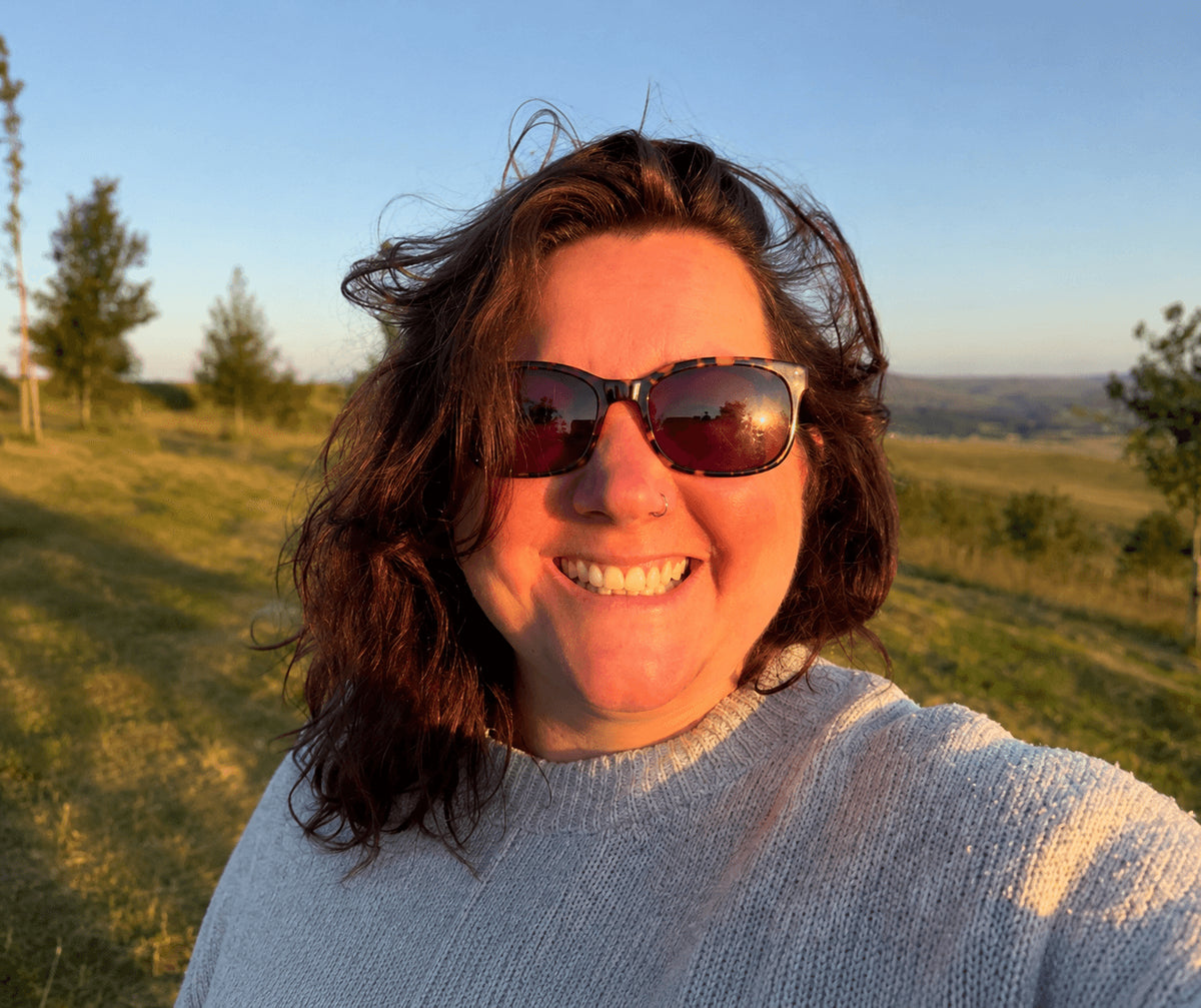 A smiling woman Francine Sharrocks Author of Wonderment - outdoors in a grassy field at sunset, wearing tortoiseshell sunglasses and a light gray knit sweater.