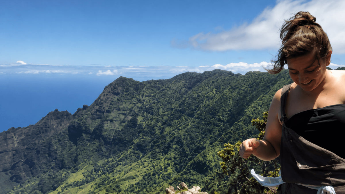 A woman in a black strapless top stands on a mountain ridge overlooking a lush green valley and ocean under a blue sky with white clouds.